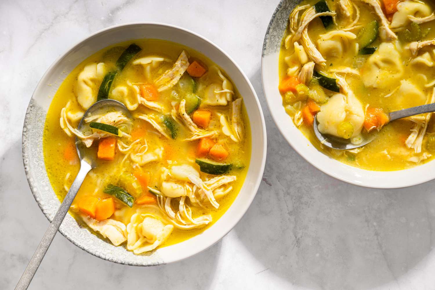 Two bowls of chicken tortellini soup with spoons containing pasta shredded chicken and diced vegetables