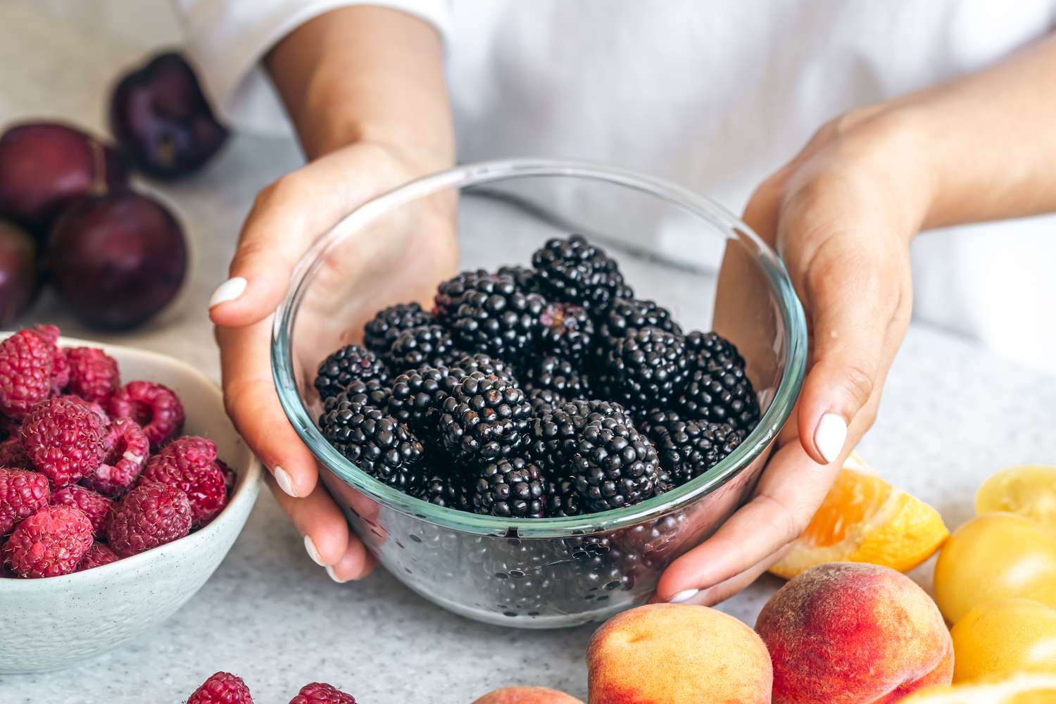 someone holding a bowl of blackberries at a table with other summer fruit