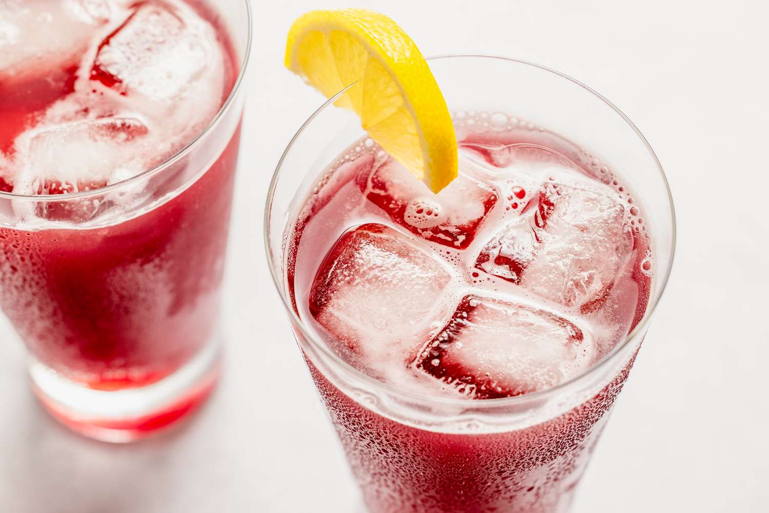 Two glasses of a red beverage with ice cubes one garnished with a lemon wedge