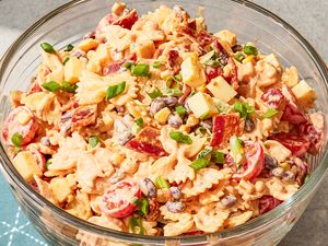 A bowl of pasta salad with tomatoes, beans, cheese, green onions, and bacon bits, on a table