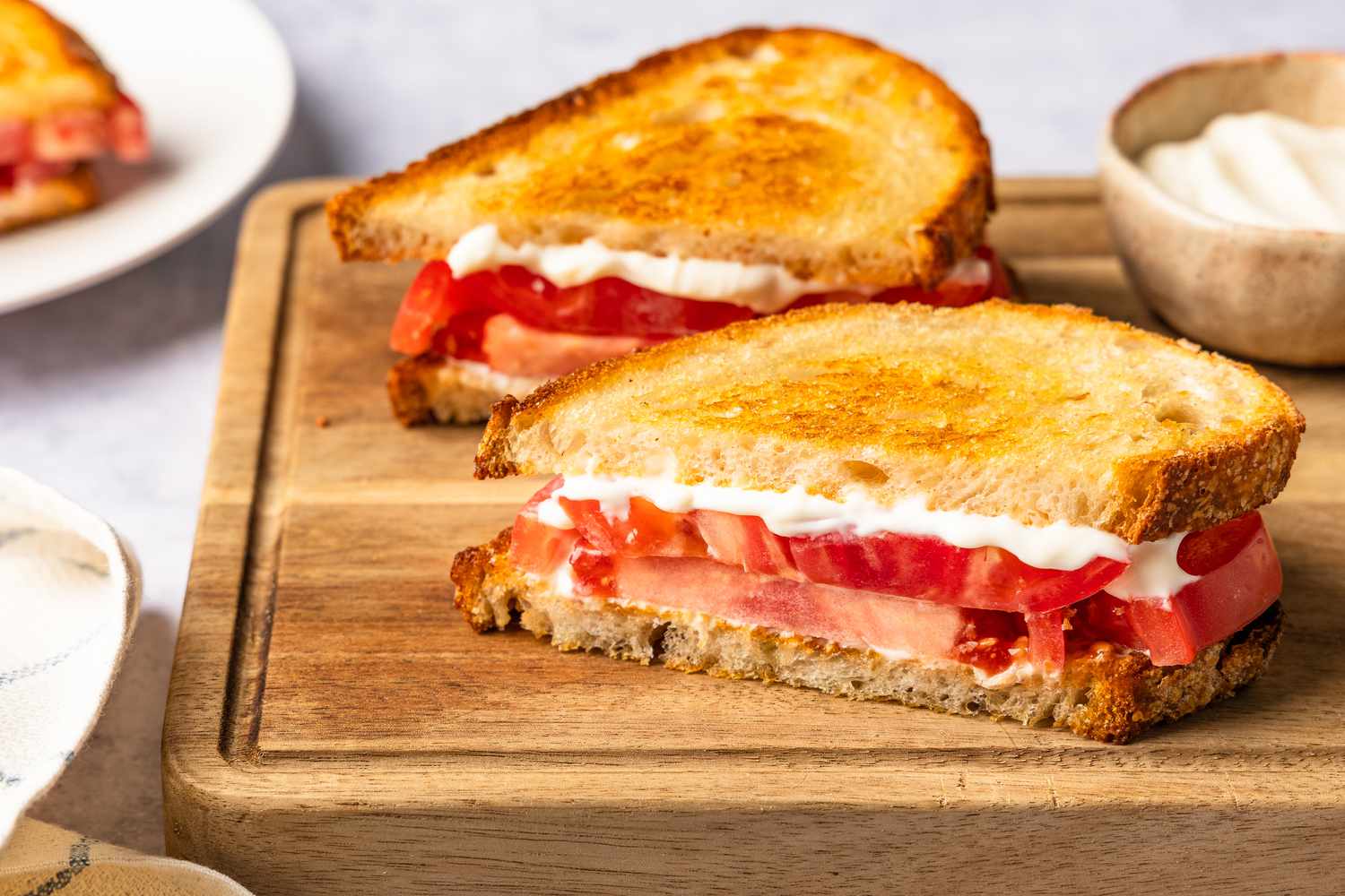 tomato sandwich halves on a cutting board next to a bowl of mayo and a plate with more tomato sandwiches