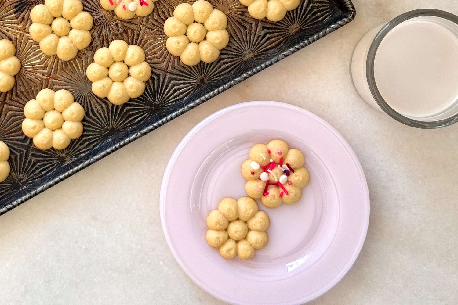 Flowershaped cookies on a baking tray next to a plate with two cookies and a glass of milk