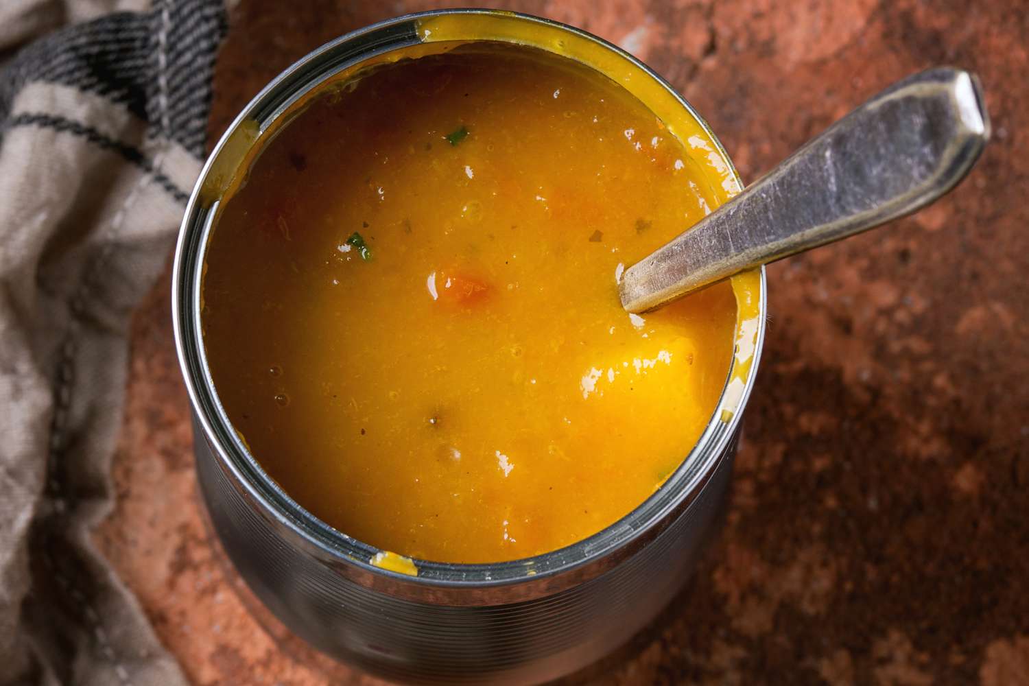 A container of orangetinted soup with a spoon inside placed on a brown surface