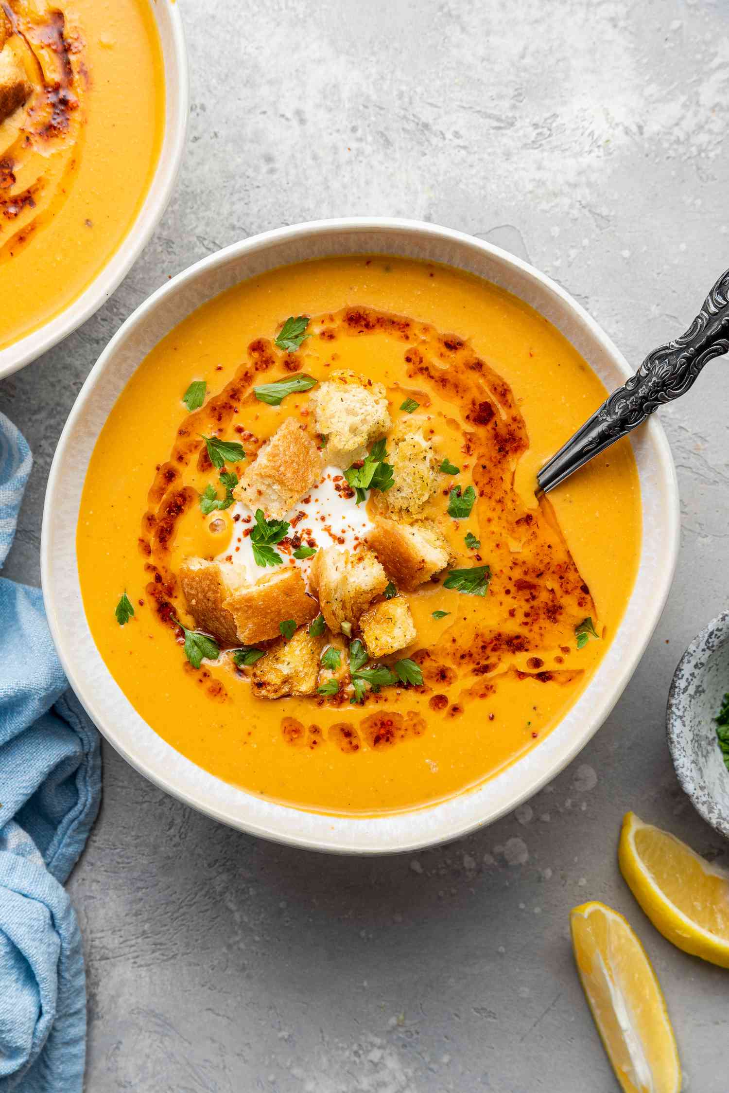 Bowl of Mercimek Corbasi (Turkish Lentil Soup) Topped with Greek Yogurt, Croutons, Pul Bibir Butter, and Parsley with a Spoon.