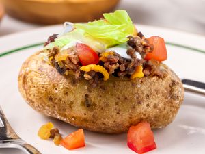 A baked potato topped with ground meat cheese tomatoes and lettuce on a plate with a fork nearby