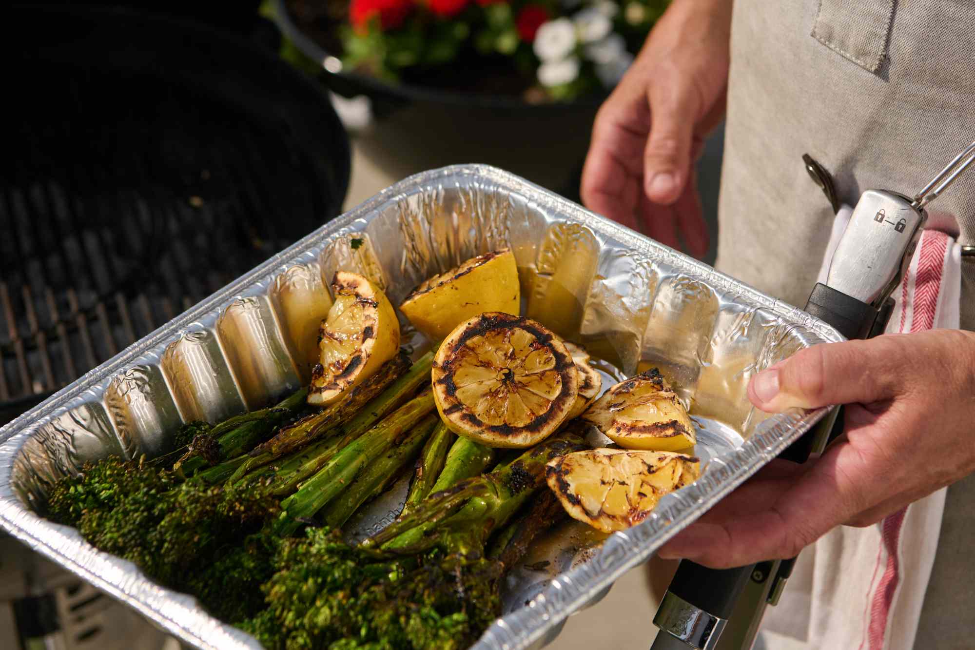 Grilled vegetables and lemon slices in a foil tray being held by someone near a grill