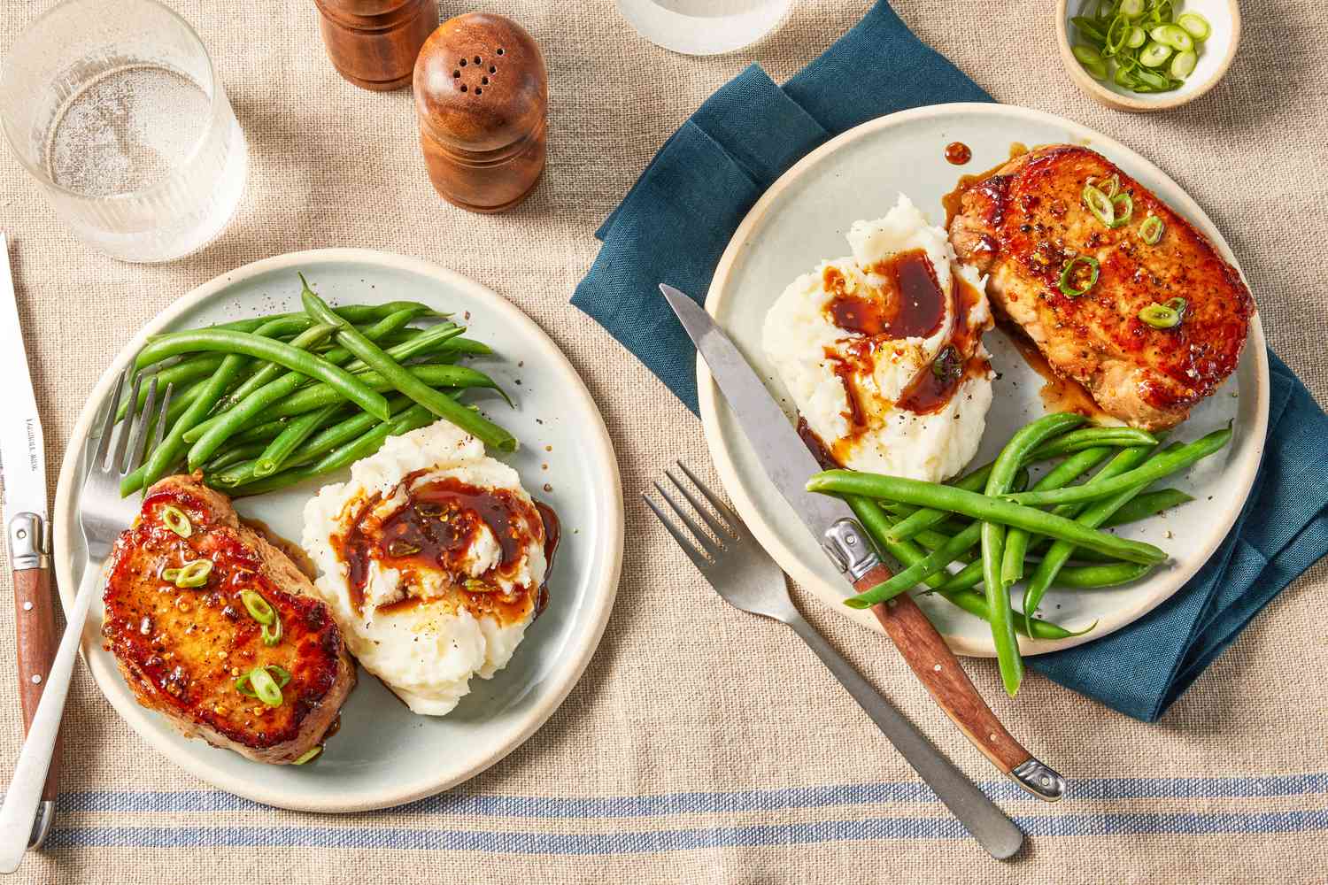 glazed pork chops served with green beans and mashed potatoes and gravy (two plates) at a table setting with utensils, a small bowl of sliced scallions, salt and pepper shakers, and a glass of water