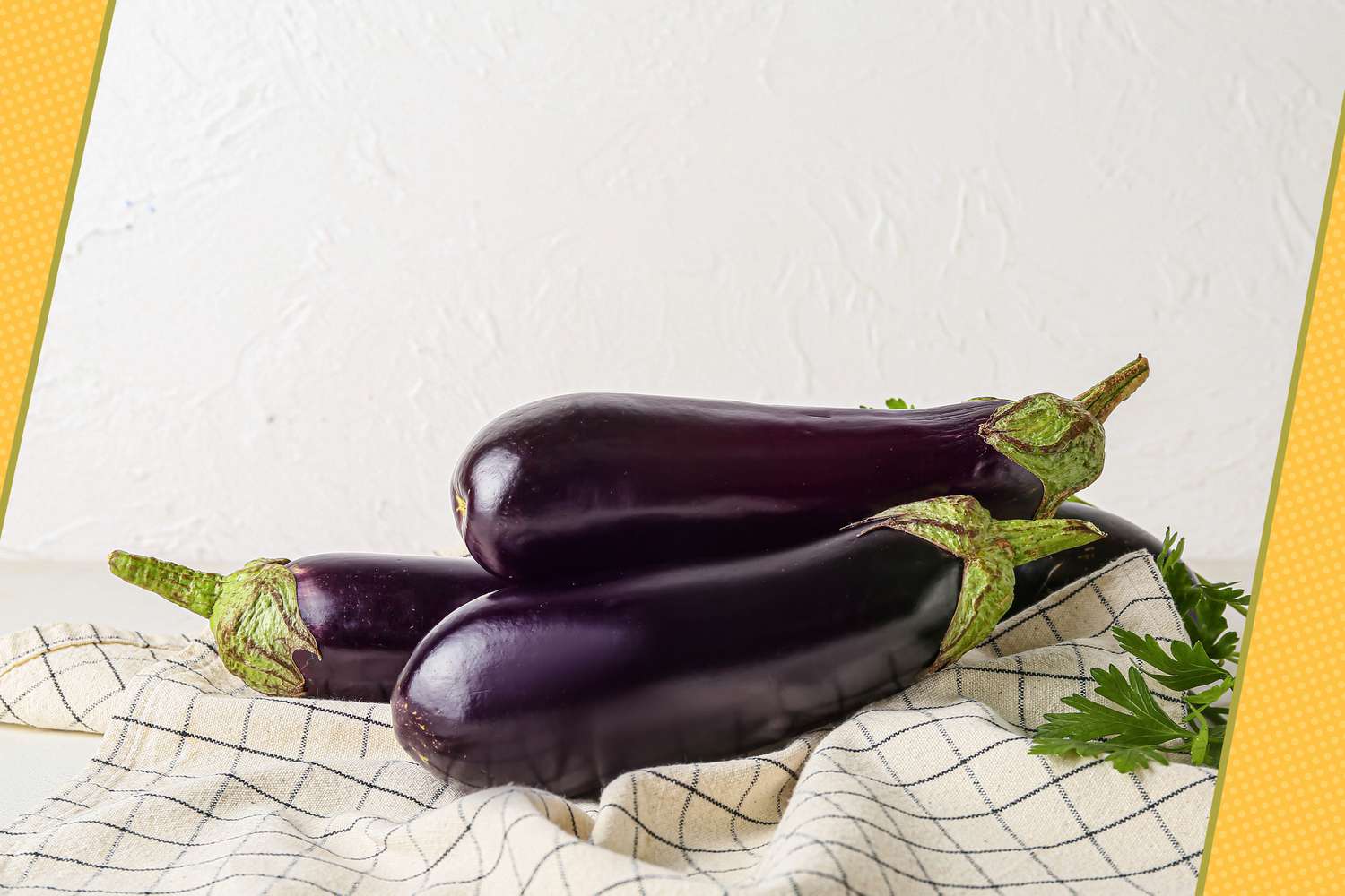 Three Eggplants Stacked Together on a White and Blue Checkered Kitchen Towel Next to Some Parsley. Edges of Photo Decorated With Yellow Polka Dotted Illustrations
