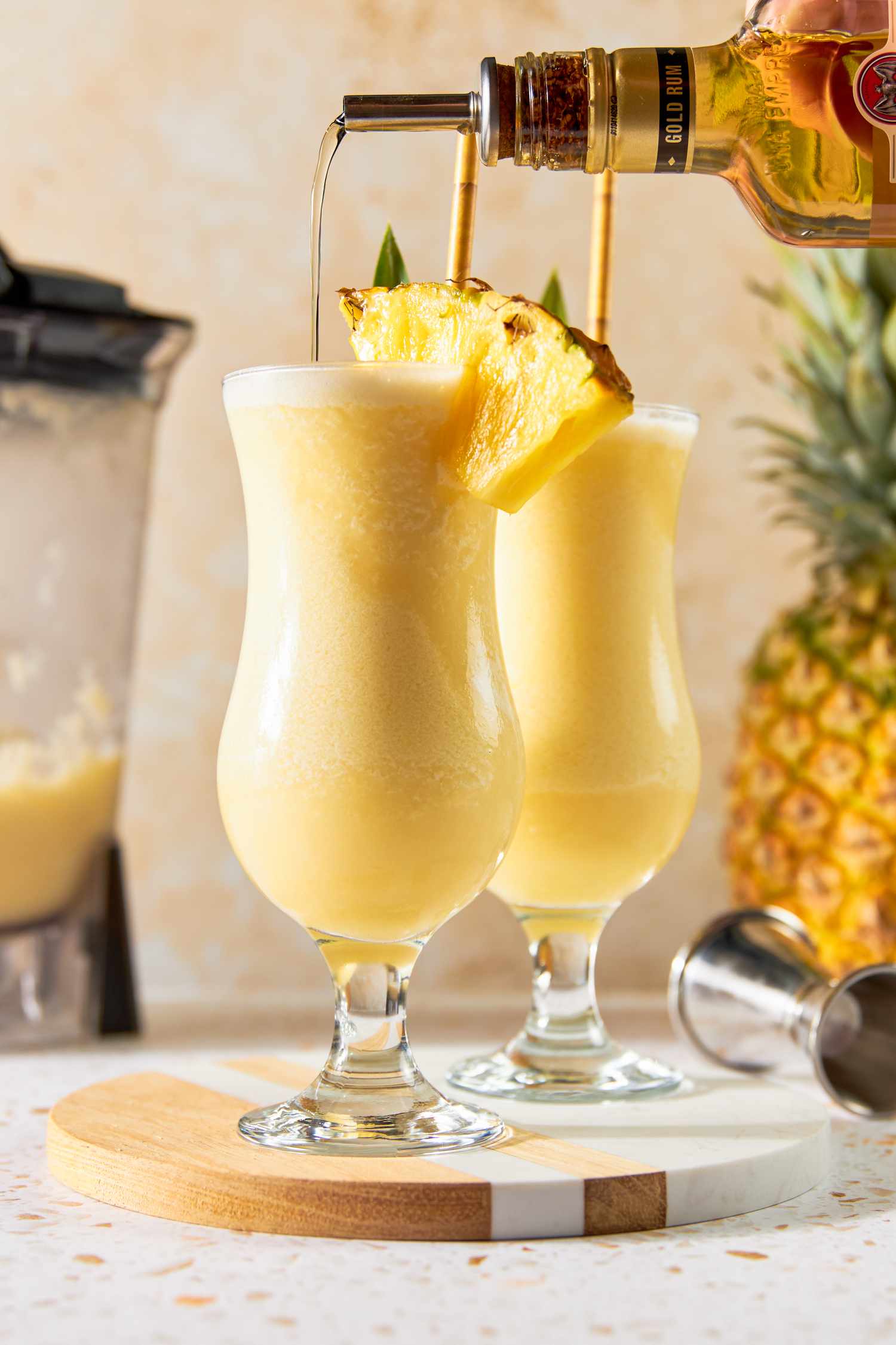 Golden Rum Poured Into a Glass of PiÃ±a Colada With a Pineapple Wedge, Pineapple Leaf, and a Straw, and Next to It, Another Serving in a Glass With the Same Garnish on a Marble and Wood Circular Serving Tray. In the Background, an Uncut Pineapple and a Blender With Some Leftover PiÃ±a Colada