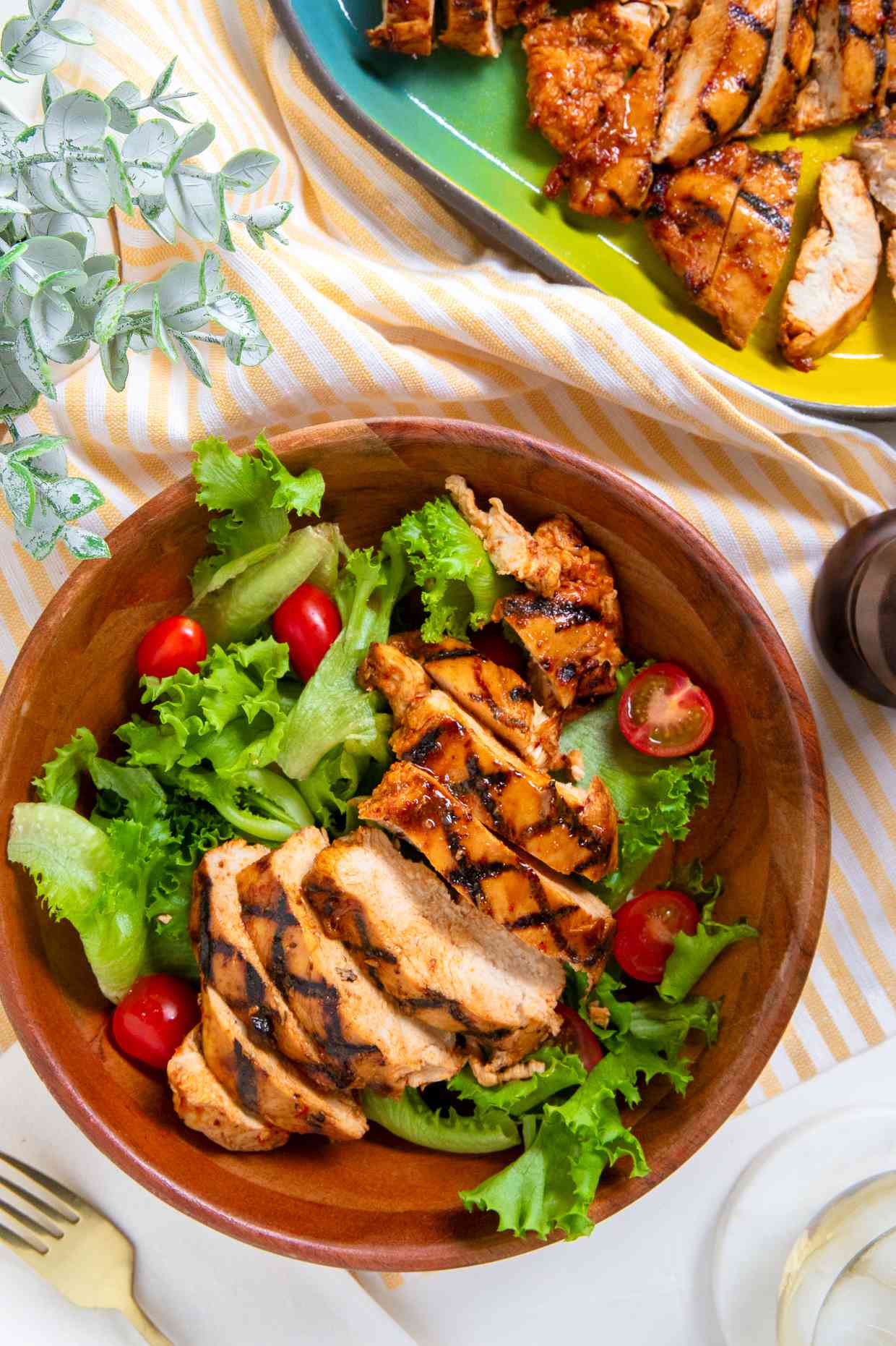 grilled chicken in a bowl over a salad at a table setting with more chicken on a platter, a glass of water, a fork, and a table plant