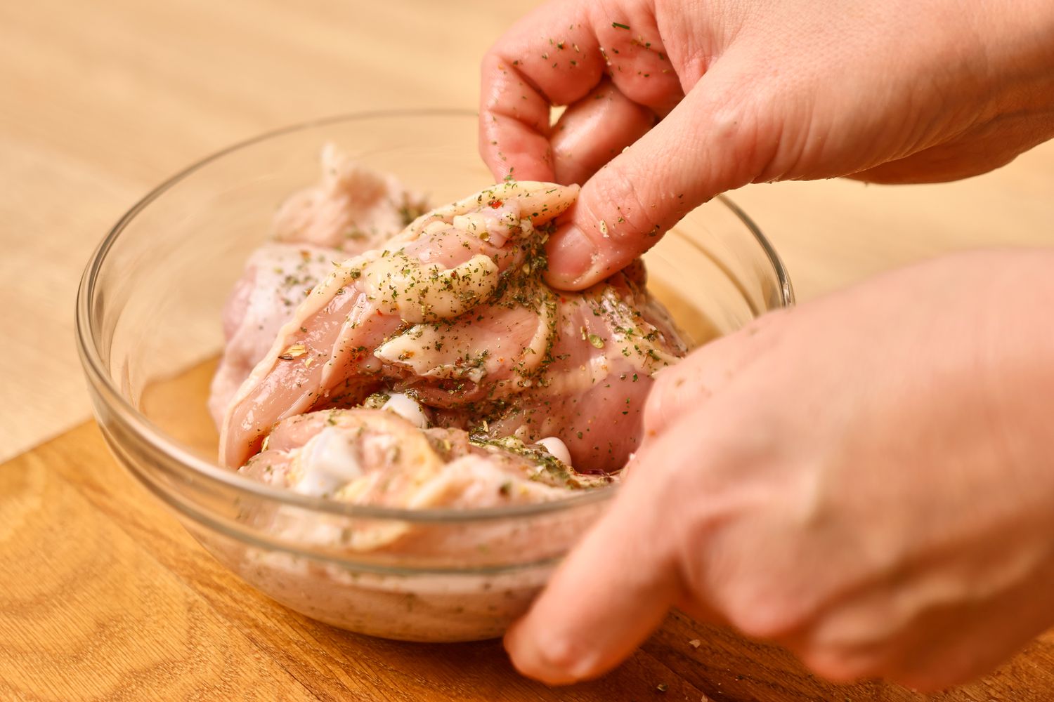 Hands dipping chicken breasts in marinade in a clear glass bowl on a wooden counter