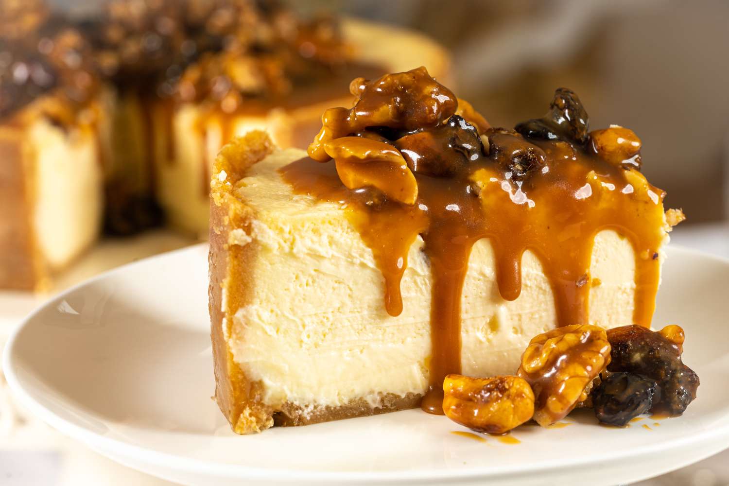 A slice of cheesecake topped with caramel and nuts on a white plate a whole cheesecake in the background