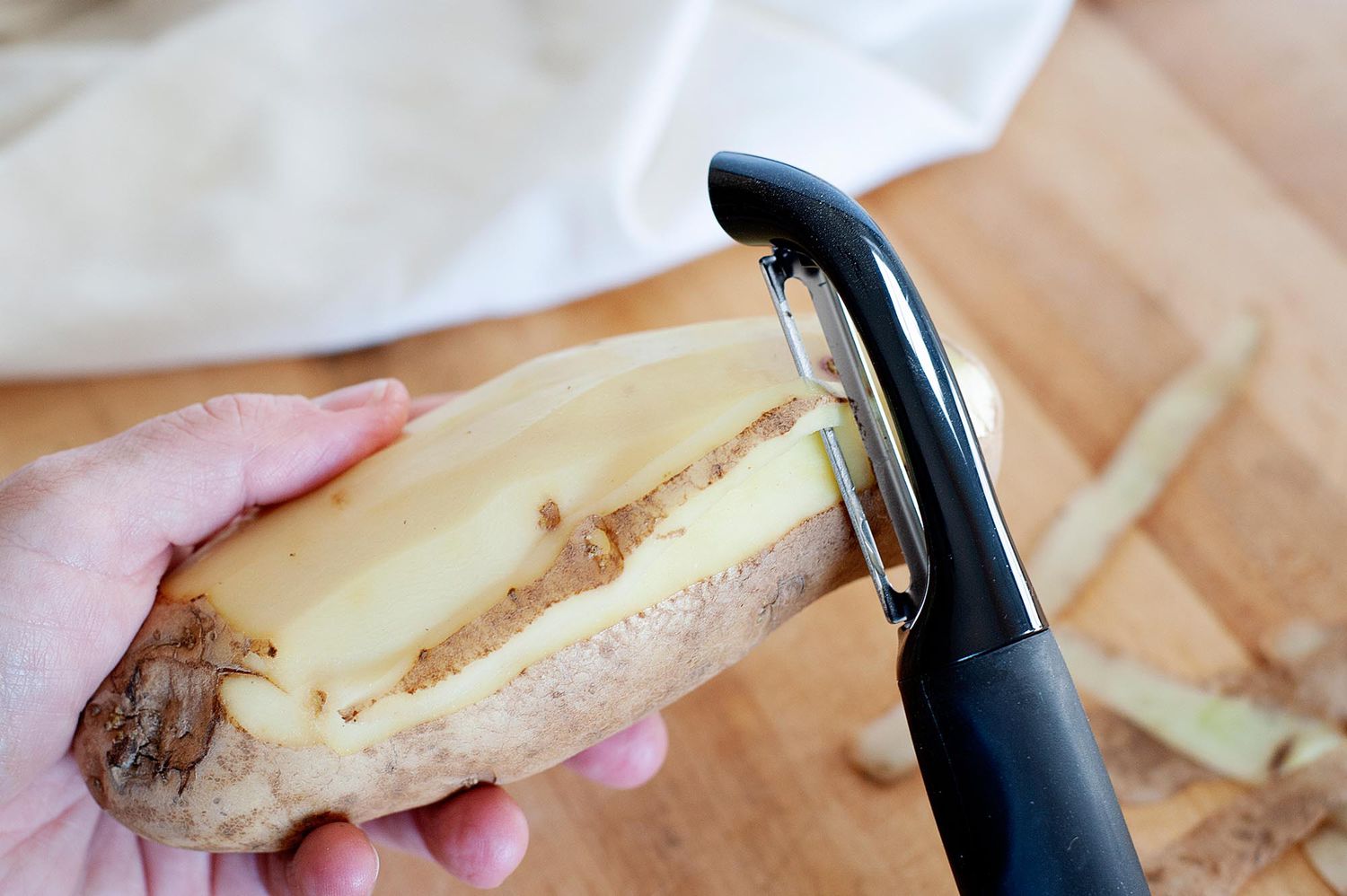 A russett potato being peeled with a black vegetable peeler.
