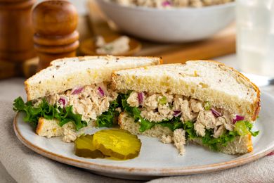 tuna salad sandwich (cut in half) on a plate with a few pickles, and in the background, more tuna salad in a bowl