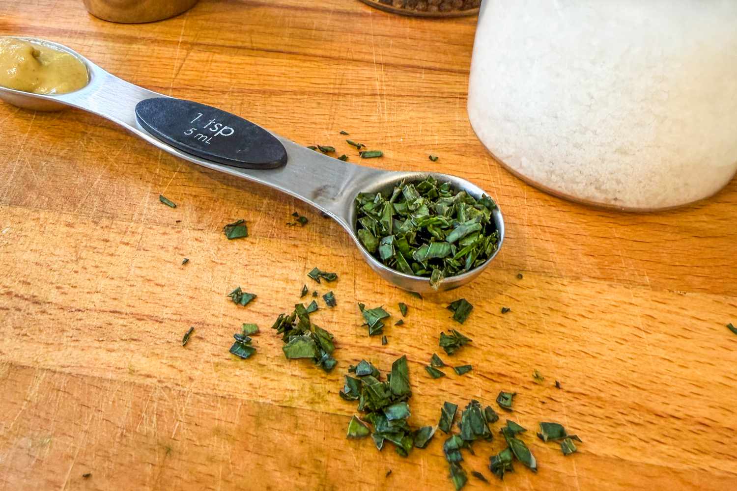 Chopped herbs on a wooden surface with a measuring spoon and a jar nearby
